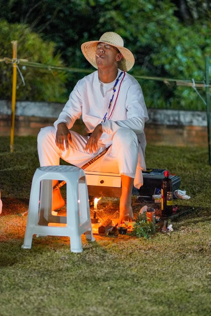 Person in white attire with hat sitting outdoors, surrounded by candles and bottles on the ground.